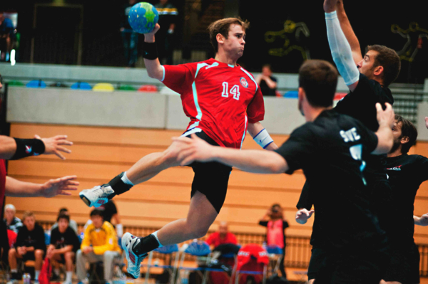 Die Männer-Handball-WM 2025 bietet Spannung und sportliche Höchstleistung! Ein männlicher Handballspieler im roten Trikot mit der Nummer 14 springt in die Luft, den blaugrünen Ball in der Hand, bereit zum Wurf, während die schwarz gekleideten Gegenspieler versuchen, ihn zu stoppen. Im Hintergrund die lebhafte Atmosphäre der Sporthalle. Hol dir dein persönliches WM-Erlebnis nachhause und sicher dir dein individuelles Tippspiel auf tippevent.de!