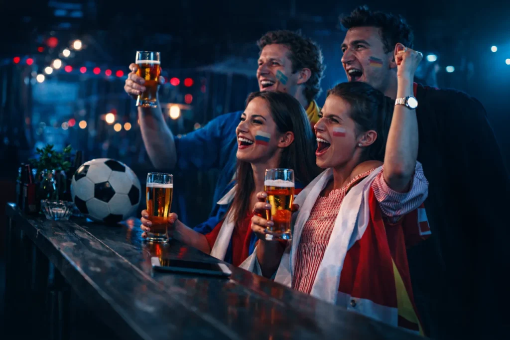 Lachende Freunde stoßen mit Bier an beim Fußball schauen in der Bar – Spaß beim Fußball-Tippspiel mit tippevent
